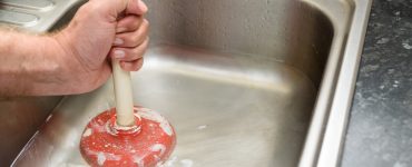 Man holding a plunger with one hand and water in sink, used to clean a clogged / blocked kitchen sink